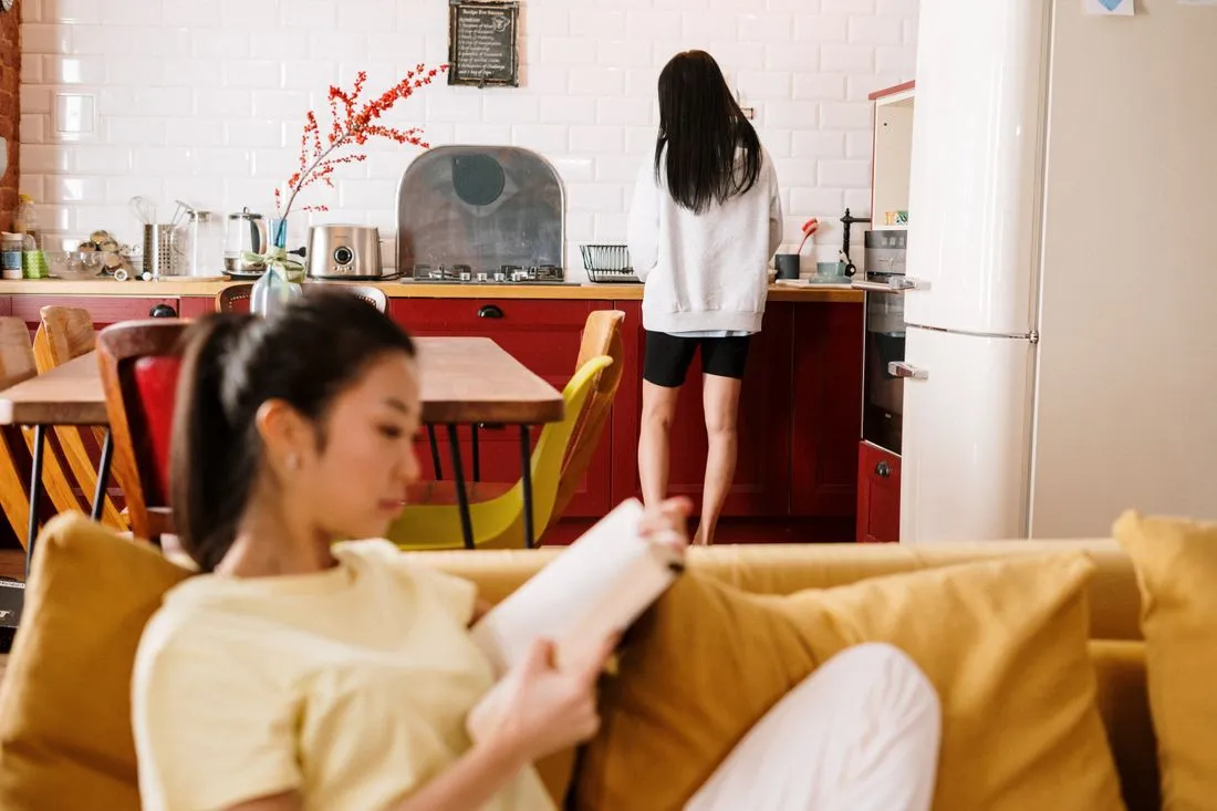 Deux jeunes femmes dans un appartement partagé, l'une lit sur le canapé, l'autre est dans la cuisine ouverte